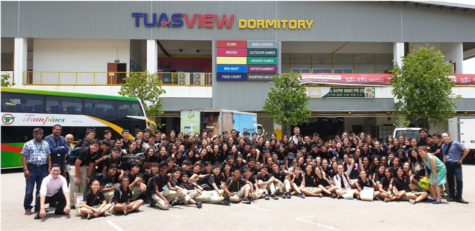 Large group of students in black uniforms posing outside Tuas View Dormitory building with a bus nearby.